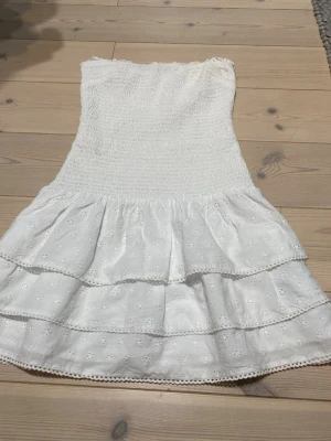Vit axelbandslös kort klänning - Söt vit axelbandslös kort klänning med smock upptill och två volanger nedtill. Klänningen har broderade detaljer och är perfekt för varma dagar. Tillverkad i bomull för en luftig och bekväm känsla. Andvänd en gång 