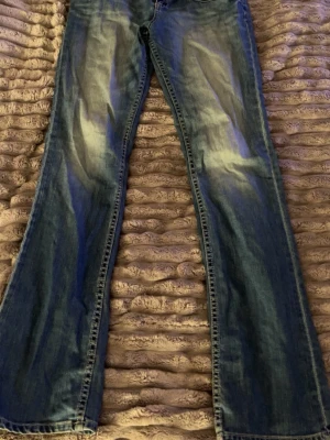 Blå bootcut jeans från Cate - Säljer ett par blå bootcut jeans från Cate med snygga slitningar och klassisk femficksmodell. Jeansen har en lätt tvättad look och är tillverkade i denim med skön stretch. Perfekta för dig som gillar en avslappnad men trendig stil.