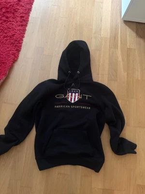 Svart hoodie från GANT med tryck - Säljer en svart hoodie från GANT med stort tryck på bröstet och texten 'American Sportswear'. Hoodien har huva med dragsko och en stor magficka. Klassisk college-look med ribbade muddar och mjukt material. Perfekt för en avslappnad stil.