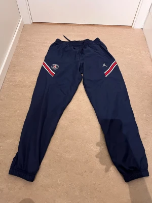 PSG träningsbyxor marinblå - Marinblå träningsbyxor från PSG x Jordan med elastisk midja och dragsko. Byxorna har röda och vita ränder vid fickorna samt PSG-logga och Jumpman-logga på framsidan. Perfekta för fotboll eller chill. Tillverkade i lätt och bekvämt material.
