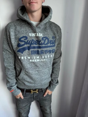 Grå Superdry hoodie med tryck - Snygg gråmelerad hoodie från Superdry med stor blå och vit logga på bröstet. Klassisk huva och känguruficka framtill. Mjuk bomullsblandning och ribbade muddar vid ärmar och midja. Perfekt för en avslappnad streetwear-look.