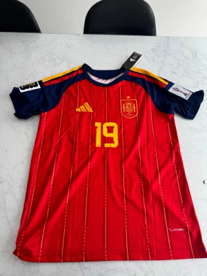 Spanien VM-Ställ Lamine Yamal nr 19  - Säljer en officiell röd matchtröja för Spaniens landslag med gula detaljer och nummer 19 på bröstet. Tröjan har blå ärmar med gula ränder och landslagets emblem på vänster sida. Tillverkad i lätt och ventilerande material.