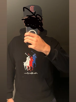 Svart hoodie med spel tryck - Säljer en svart hoodie med stor ficka framtill och huva. På bröstet finns ett färgglatt tryck med tre polospelare i vitt, rött och blått. Hoodien har en avslappnad passform och är perfekt för en chill look. Stretcit material. Är storlek xl men enligt mig så är den liten så typ en L! Obs liten i storlek. Mer som L. Tvätt lappen är tyvär borta