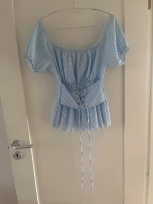 Blå rutig blus med snörning - Supersöt blå och vit rutig blus med puffärmar och snörning i midjan. Toppen har offshoulder-modell, smock i ryggen och volang nedtill. 