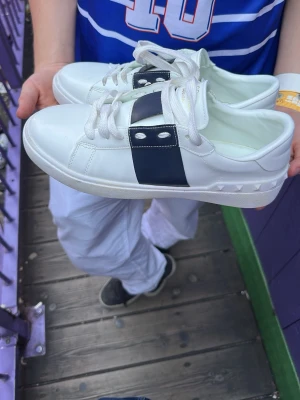Valentino garavani - Snygga vita sneakers från Valentino Garavani med bred svart rem och nitar på sidan. Skorna har vita skosnören och en klassisk låg profil. Tillverkade i slätt läder med gummisula och coola detaljer som ger en exklusiv vibe.