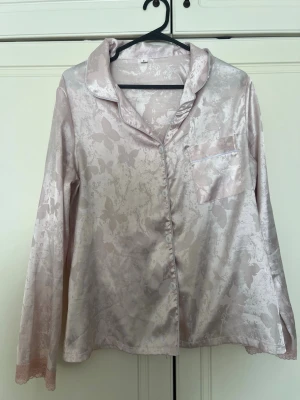 Ljusrosa satinpyjamas med fjärilsmönster - Snygg ljusrosa pyjamaströja i glansigt satinmaterial med subtilt fjärilsmönster. Skjortmodell med krage, knappstängning och en bröstficka. Perfekt för mysiga kvällar hemma.