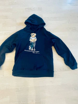 Svart hoodie Polo Ralph Lauren - Snygg svart hoodie från Polo Ralph Lauren med den klassiska björnen på bröstet. Hoodien har huva och stor magficka framtill. Tillverkad i mjukt material som känns skönt mot huden. Perfekt för dig som gillar streetwear och vill ha en ikonisk look.