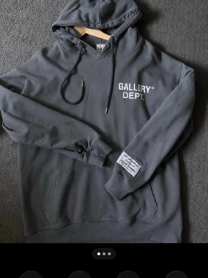 Mörkgrå hoodie Gallery Dept. - Snygg mörkgrå hoodie från Gallery Dept. med vit logga tryckt på bröstet och stor text på ryggen. Klassisk känguruficka framtill, ribbade muddar och justerbar huva med snören. Liten patch på ärmen för extra detalj. Perfekt för en avslappnad streetwear-look.