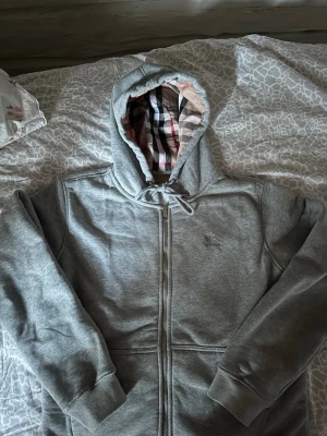 En riktig snygg BB Hoodie - Snygg grå zip-hoodie med huva och dragsko. Insidan av huvan och öppningen har ett klassiskt rutigt mönster i beige, svart, vitt och rött. Hoodien har långa ärmar, ribbade muddar och två fickor framtill. Perfekt för en chill och stilren look.  Bara testat denna Men tyckte den ej passade mig, gick runt med denna kanske 20-40 minuter annars helt sprillans ny😊