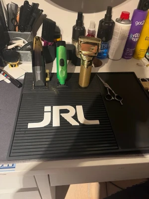 JRL klippmaskin och frisörkit - Säljer ett komplett frisörkit med JRL klippmaskin i svart och guld, grön trimmer, guldfärgad rakapparat samt sax. Perfekt för dig som vill fixa snygga frisyrer hemma. Allt ligger på en svart JRL matta och är redo att användas.