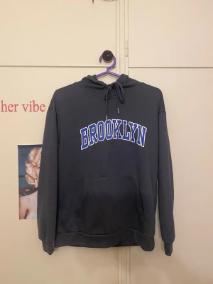 Mörkgrå hoodie med BROOKLYN-tryck - Snygg mörkgrå hoodie med stor vit och blå BROOKLYN-text på bröstet. Klassisk känguruficka framtill, huva med dragsko och ribbade muddar vid ärmar och nederkant. Perfekt för en chill och avslappnad stil.