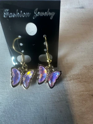 Butterfly Earrings on Display Card - This image shows a pair of butterfly-shaped earrings with a shiny, iridescent purple finish. The earrings are hanging on a black display card labeled 'Fashion Jewelry.' The hooks and earring frames appear to be gold-toned. The background is a light-colored textured surface, and a yellow measuring tape is partially visible in the upper left corner of the second image.
