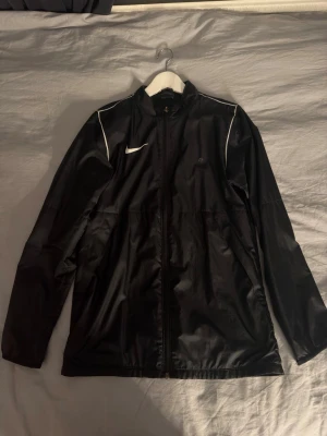 Svart träningsjacka från Nike - Snygg svart träningsjacka från Nike med hel dragkedja framtill och vita detaljer på axlarna. Jackan har hög krage, långa ärmar och är tillverkad i ett lätt, sportigt material som passar perfekt till träning eller uppvärmning.