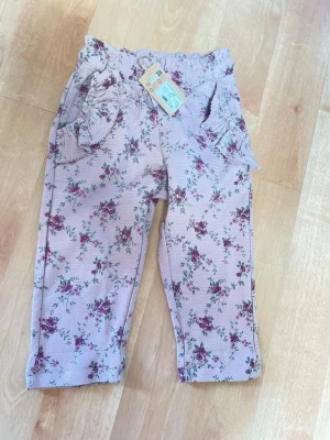 Blommiga leggings med volangfickor,92 - Supersöta leggings i ljusrosa med blommigt mönster i lila och grönt. De har volangdetaljer på fickorna framtill och en bakficka med liten etikett. Mjuk resår i midjan och stretchigt material för skön passform.