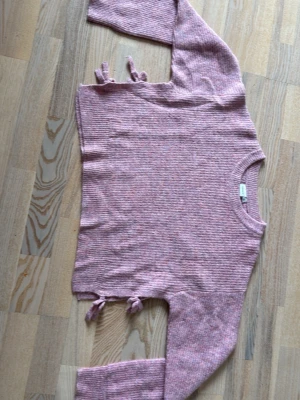 Rosa stickad tröja från Name It - Mysig rosa stickad tröja från Name It med ribbad struktur och dekorativa knytband i sidorna. Tröjan har rund halsringning och långa ärmar. Perfekt för dig som gillar en avslappnad och trendig stil.