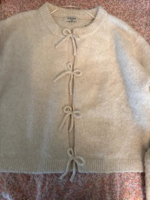 Vit kofta med rosettknäppning SAMSA - Supersöt vit kofta från SAMSA med fyra dekorativa rosetter som knäppning framtill. Koftan är mjuk och fluffig med ribbade muddar vid hals, ärmslut och nederkant. Perfekt för dig som gillar en gullig och trendig stil.