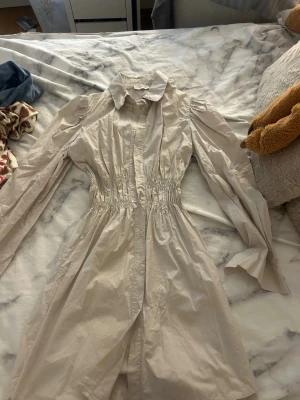 Ljusbeige skjortklänning med lång ärm - Säljer en ljusbeige skjortklänning med markerad midja och långa ärmar. Klänningen har krage, knappar framtill och puffiga axlar för en trendig look. Perfekt för dig som gillar stilrena och klassiska plagg.