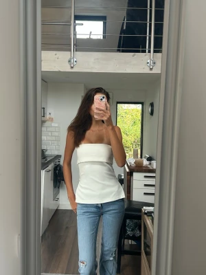 Vit axelbandslös tubtopp - Säljer en stilren vit tubtopp med figurnära passform och axelbandslös design. Toppen har en enkel och clean look som passar perfekt till jeans eller kjol. Dragkedjor på sidorna.