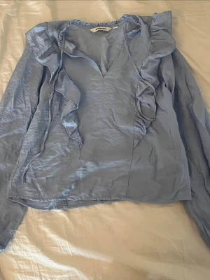 Ljusblå volangblus från Kappahl - Supersöt ljusblå blus från Kappahl med långa ärmar och volangdetaljer längs v-ringningen och framtill. Tillverkad i ett tunt, lätt material som ger en luftig känsla. Perfekt för dig som gillar romantiska detaljer och en feminin look.