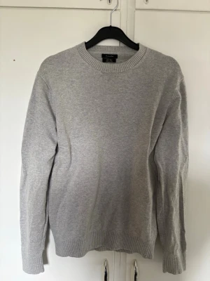 Ljusgrå stickad tröja från Massimo Dutti - En stilren ljusgrå stickad tröja med rund halsringning och ribbade muddar vid ärmslut och nederkant. Perfekt för lager på lager och enkel att matcha med olika outfits. Tröjan har en klassisk passform och är mjuk i materialet. Bra skick, men litet hål runt halsen 