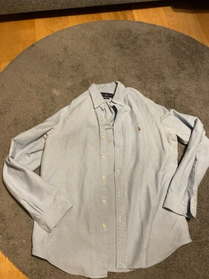 Ljusblå skjorta från Ralph Lauren - Klassisk ljusblå skjorta från Ralph Lauren med button-down krage och broderad logga på bröstet. Skjortan har långa ärmar, normal passform och stängs med vita knappar framtill. Perfekt till jeans eller chinos för en clean look.