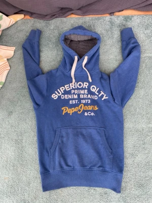 Blå hoodie från Pepe Jeans - Säljer en blå hoodie från Pepe Jeans med vit och gul text på bröstet. Tröjan har huva med vita snören, magficka och mjukt grått foder i luvan. Perfekt för dig som gillar archive och vill ha något bekvämt och snyggt.