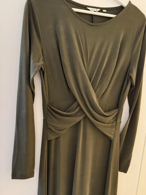 Midiklänning - Snygg mörk olivgrön midiklänning med omlottdetalj framtill och långa ärmar. Klänningen har en mjuk, följsam känsla och rund halsringning. Perfekt för dig som gillar stilrena och eleganta plagg med en twist.