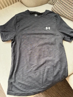 Under Armour tränings t-shirt – M - Mörkgrå/svart tränings t-shirt från Under Armour i storlek M. Fitted passform och lätt, funktionellt material som passar perfekt för gym, löpning och annan träning. Under Armour-logga framtill samt större logga upptill på ryggen.  Fint begagnat skick.