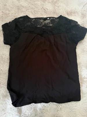 Svart topp med spetsdetaljer - Snygg svart topp med korta ärmar och transparent spets upptill samt över axlarna. Toppen har rund halsringning och ett enkelt, rakt snitt som passar till många olika stilar. Perfekt för dig som gillar en stilren look med lite extra detalj. Det är inte en röd stor cirkel i mitten utan det blev av kamran.