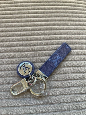 Vuitton Blå nyckelring med metallhänge - Snygg Vuitton  blå nyckelring med ett rektangulärt band i präglat läder och en rund metallbricka med bokstäver. Nyckelringen har både ring och karbinhake i silverfärgad metall, perfekt att fästa på väskan eller nycklarna.