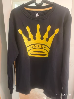 Svart stickad tröja med gul krona - Cool svart stickad tröja från MR med en stor gul krona och texten 'MR' på bröstet. Tröjan har rund halsringning och långa ärmar. Perfekt för dig som vill sticka ut med en unik och trendig look.