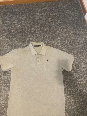 Ljusgrå pikétröja med krage - Snygg ljusgrå pikétröja med klassisk krage och knappar framtill. Tröjan har korta ärmar och en liten broderad logga på bröstet. Perfekt till jeans eller shorts för en clean och enkel stil.