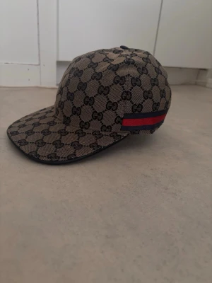 Gucci beige keps med GG-mönster - Säljer en beige keps från Gucci med klassiskt GG-monogram över hela kepsen och en röd-blå rand på sidan. Kepsen har böjd skärm och justerbar rem bak. Materialet ser ut att vara canvas med detaljer i tyg. Perfekt för dig som gillar exklusiva accessoarer.