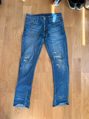 Blå slitna jeans från Nudie Jeans - Säljer ett par blå jeans från Nudie Jeans med snygga slitningar och tvättade detaljer. Klassisk femficksmodell med raka ben och normal midja. Jeansen har Nudie Jeans signaturbroderi på bakfickan och är gjorda i denim av bomull.