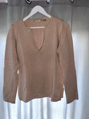 Beige stickad tröja med v-ringning - Snygg beige stickad tröja med djup v-ringning och långa ärmar. Tröjan har en enkel, clean design och ribbade kanter vid halsen. Perfekt att styla med jeans eller kjol för en avslappnad look.