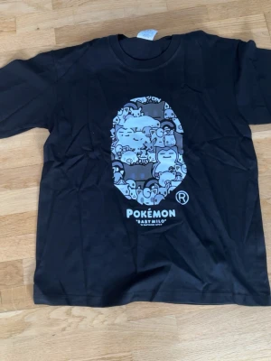 Bape x Pokemon T-shirt  - Svart T-shirt med Pokémon x Baby Milo-tryck i blå toner på bröstet. Motiv med flera Pokémon-karaktärer i en ikonisk Baby Milo-stil. Rund hals och korta ärmar. Perfekt för dig som gillar streetwear och anime.