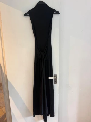 Svart långklänning med knyt - Stilren svart långklänning med höghalsad design och snygg knytning i midjan. Klänningen har ärmlös modell och ett mjukt, följsamt tyg som ger en elegant siluett. Perfekt för dig som gillar minimalistisk och chic stil.
