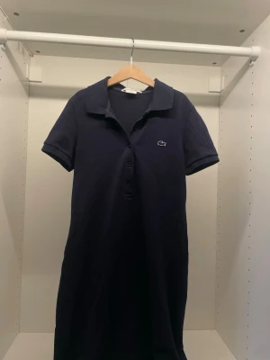 Mörkblå pikéklänning från Lacoste - Klassisk mörkblå pikéklänning från Lacoste med korta ärmar, krage och knappar framtill. Klänningen har den ikoniska krokodilloggan på bröstet och är gjord i ett mjukt pikématerial som känns sportigt och stilrent.