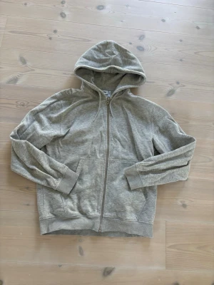Ljusgrå zip hoodie med huva Weekday - En stilren ljusgrå hoodie med dragkedja framtill och två fickor. Hoodien har en klassisk huva och ribbade muddar vid ärmar och nederkant. Perfekt för en avslappnad look och enkel att matcha med andra plagg.