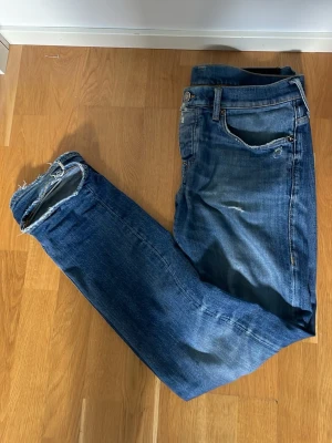 Blå True Religion jeans med slitningar - Säljer ett par blå jeans från True Religion, modell Rocco, med klassiska bakfickor och slitna detaljer vid benslut och fickor. Jeansen har en snygg tvätt och raka ben. Perfekta för dig som gillar en avslappnad och trendig stil.                PRIS KAN DISKUTERAS