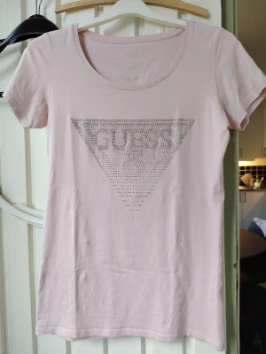 Ljusrosa t-shirt med strassdetaljer - Snygg ljusrosa t-shirt med korta ärmar och rund halsringning. Framtill finns ett stort triangelformat tryck med strassdetaljer som bildar texten 'GUESS'. Perfekt till jeans eller kjol för en trendig look.