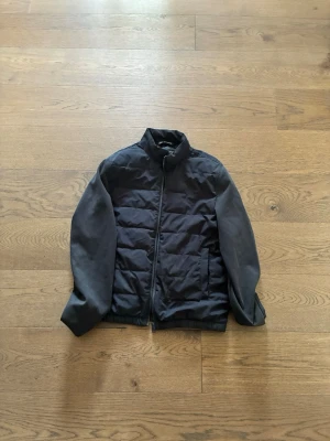 Svart pufferjacka med ståkrage - Säljer en svart pufferjacka med ståkrage och dragkedja framtill. Jackan har quiltade partier på kroppen och släta ärmar. Två fickor framtill och elastisk nederkant. Perfekt för kalla vinterdagar och riktigt skön att bära.