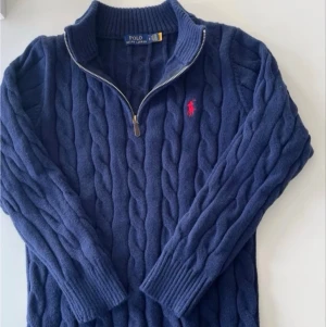 Mörkblå kabelstickad tröja Polo Ralph Lauren - Kabelstickad mörkblå tröja från Polo Ralph Lauren med halv dragkedja och ribbade muddar. Klassisk röd logga broderad på bröstet. Perfekt för lager på lager och riktigt snygg till jeans.