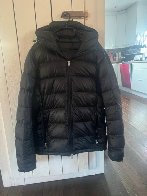 Svart J. Lindeberg pufferjacka - Säljer en svart J. Lindeberg pufferjacka med huva och dragkedja framtill. Jackan har två fickor med dragkedja och quiltad design som håller värmen bra. Perfekt för kalla dagar och har en klassisk, stilren look. Jackan har ett litet hål på baksidan men det är under huvan så man ser det oftast inte. En av muddarna är också lite nött (se bild). Priset är inte hugget i sten