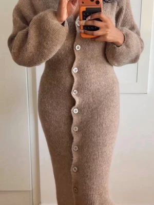 Beige stickad långklänning - Mysig beige stickad långklänning med långa ärmar och dekorativa vita knappar framtill. Klänningen har en figurnära passform och är perfekt för kyligare dagar. Den ribbade stickningen ger en mjuk och bekväm känsla.