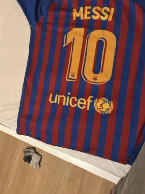 Barcelona Messi matchtröja blå/röd - Säljer en officiell FC Barcelona matchtröja med Messi och nummer 10 på ryggen. Tröjan är blå och röd med vertikala ränder, har klubbmärke och Nike-logga på bröstet samt Rakuten och Unicef-tryck. Tillverkad i lätt och ventilerande material, perfekt för fotboll.