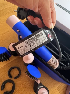 Dyson blå hårstyler med tillbehör - Blå och rosa hårstyler från Dyson med flera utbytbara borsthuvuden och tillbehör. Cylindrisk form, EU-kontakt och tillverkad i plast och metall. Perfekt för att fixa olika frisyrer och styling. Kommer med praktisk förvaringsväska.