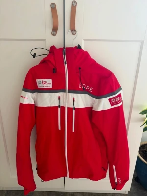 Röd vindjacka EQPE Åre 2019 - Snygg röd vindjacka från EQPE med vita och grå detaljer, officiell jacka från Åre 2019. Jackan har huva, hel dragkedja, flera fickor med dragkedja och tryckta loggor på bröst, ärm och rygg. Perfekt för dig som gillar sportig stil.