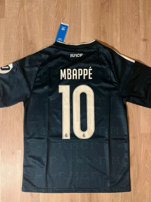 Real Madrid Mbappé Bortatröja 26/27 Mörkgrön - Snygg mörkgrön Real Madrid matchtröja med Mbappé och nummer 10 på ryggen. Tröjan har vita detaljer, klubbmärke på bröstet samt sponsortryck framtill. Tillverkad i lätt och ventilerande material, perfekt för fotbollsträning. HELT NY!