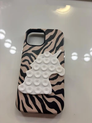 Zebramönstrat mobilskal med octo Buddy - Snyggt mobilskal med zebramönster i beige och svart. På baksidan sitter en vit octobuddy i form av en enhörning, perfekt för dig som gillar att pilla på något. Till iPhone 15